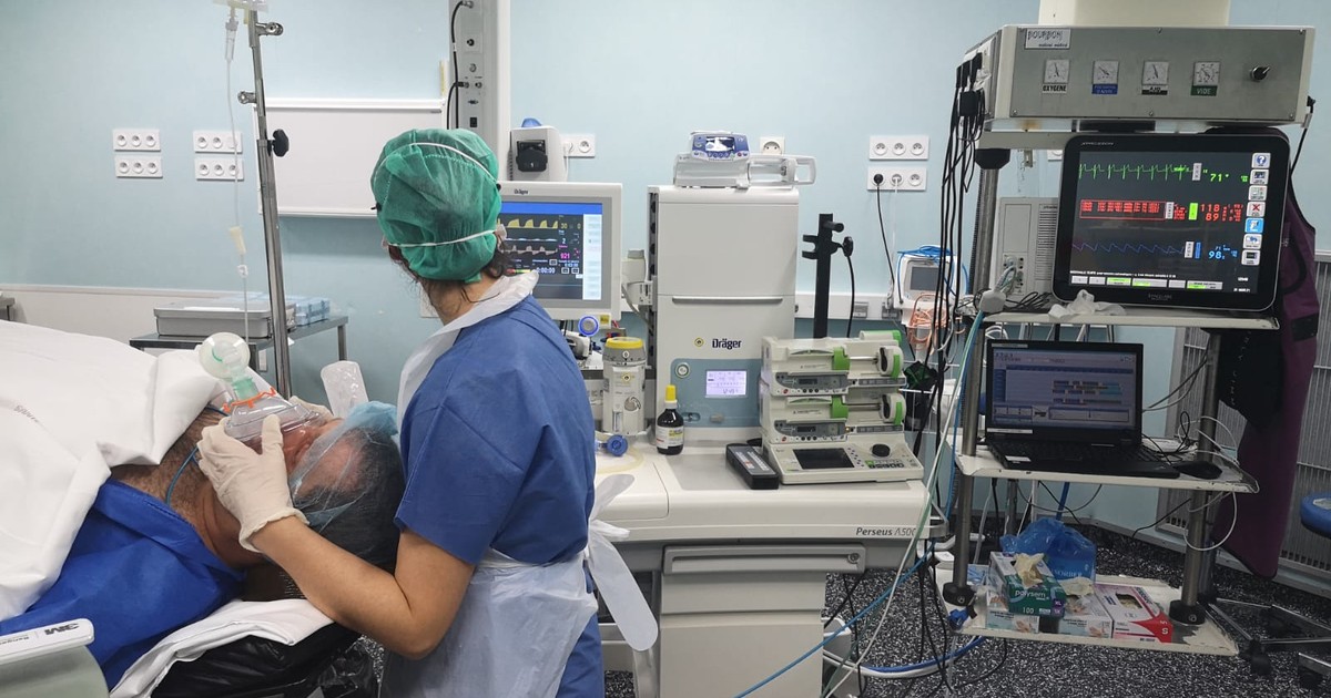 An anesthetist holds a mask over a patient's face at the start of anesthesia, with the anesthesia machine and vital-signs monitors beside them in an operating room